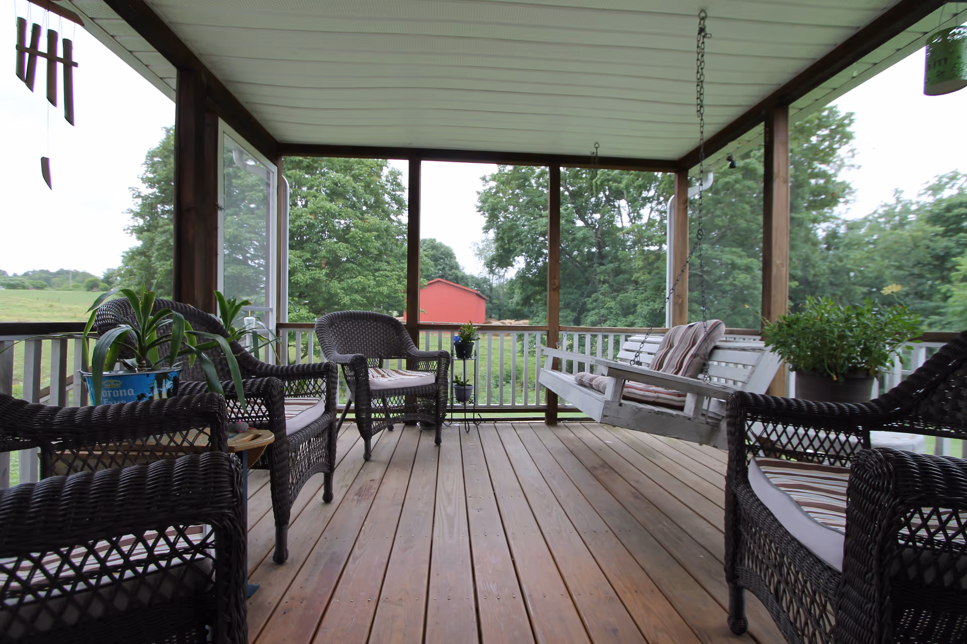 A covered wooden porch with several wicker chairs and a white wooden swing with cushions. There are potted plants on the porch and a view of green trees and a red barn in the background.