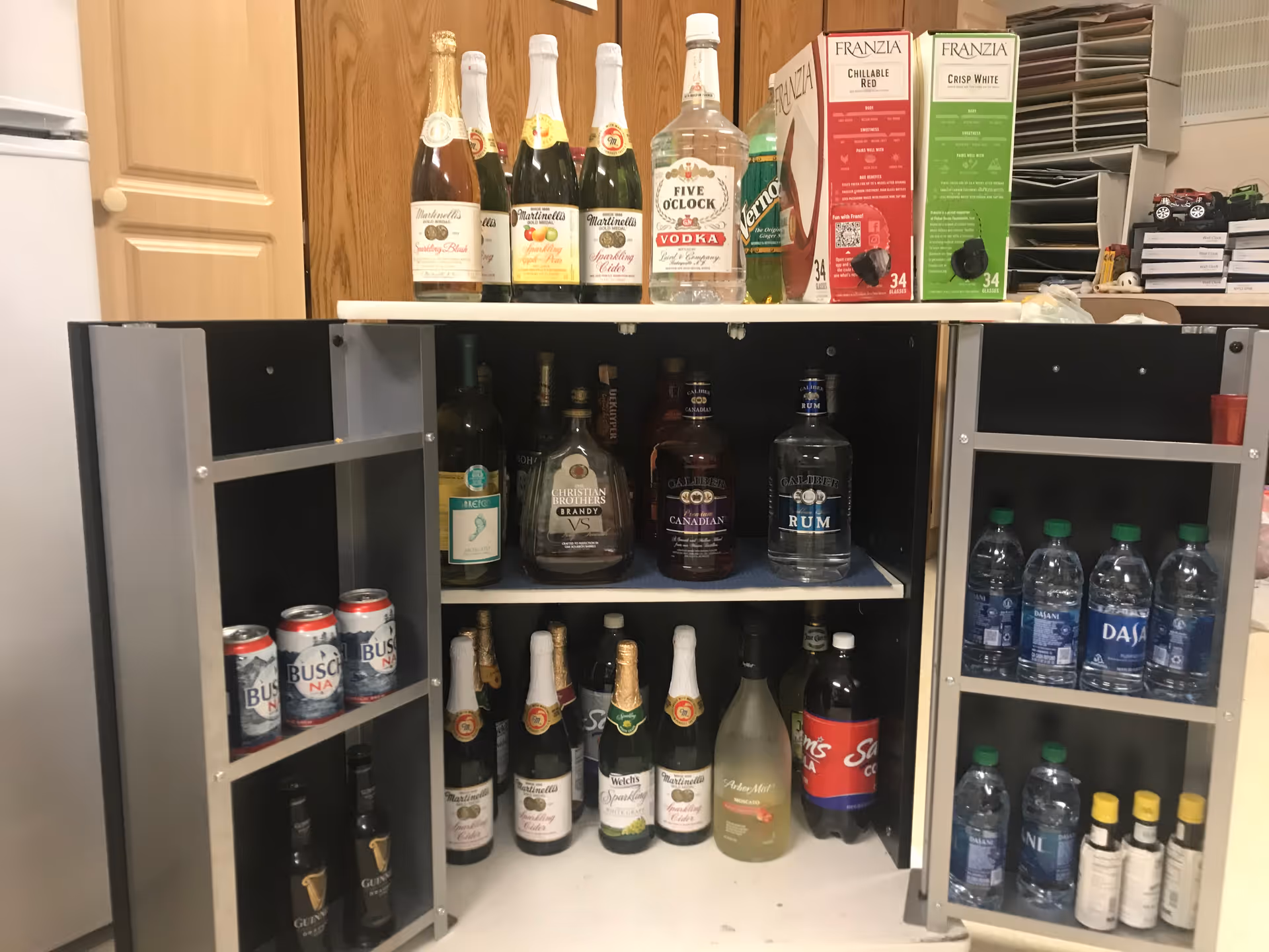 Open cabinet with various alcoholic beverages and bottled water. The top shelf holds bottles of sparkling cider, vodka, and boxed wine. The middle shelf contains bottles of brandy, Canadian whiskey, and rum. The bottom shelf has more sparkling cider bottles, a bottle of white wine, and a soda bottle. The cabinet doors have shelves stocked with cans of non-alcoholic beer, bottles of Guinness, bottled water, and small bottles of liquor or mixers.