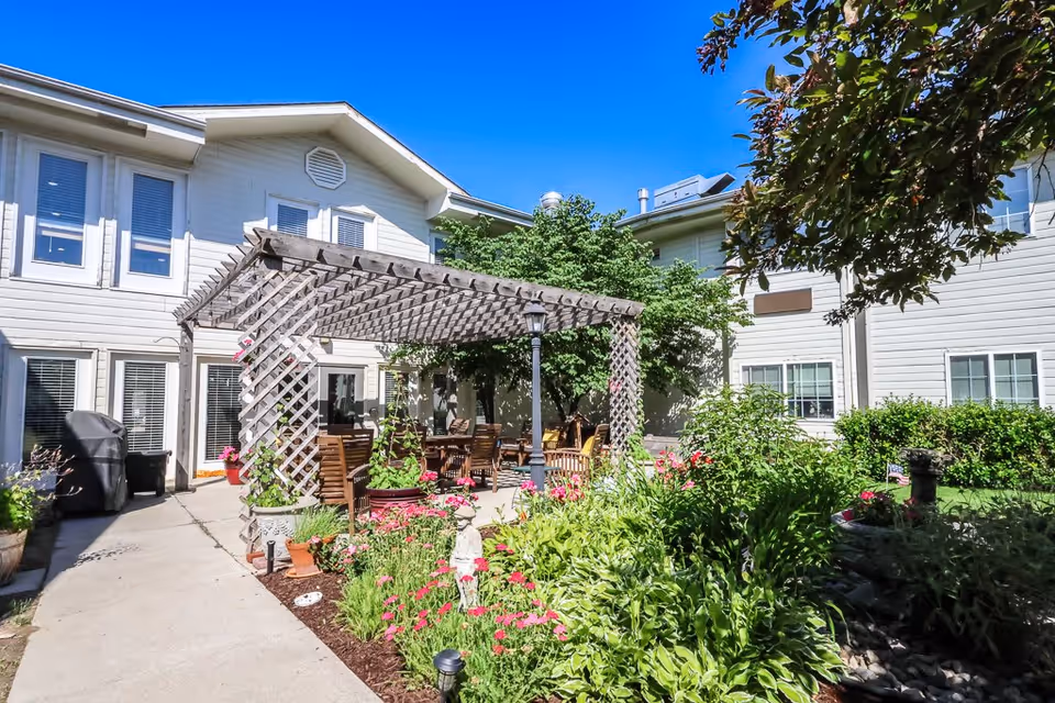 Outdoor patio area at Bailey Pointe at Miracle Hills featuring a wooden pergola with climbing plants, wooden chairs and tables underneath, surrounded by lush green plants and colorful flowers. The building exterior is light-colored with multiple windows, and the sky is clear and blue.