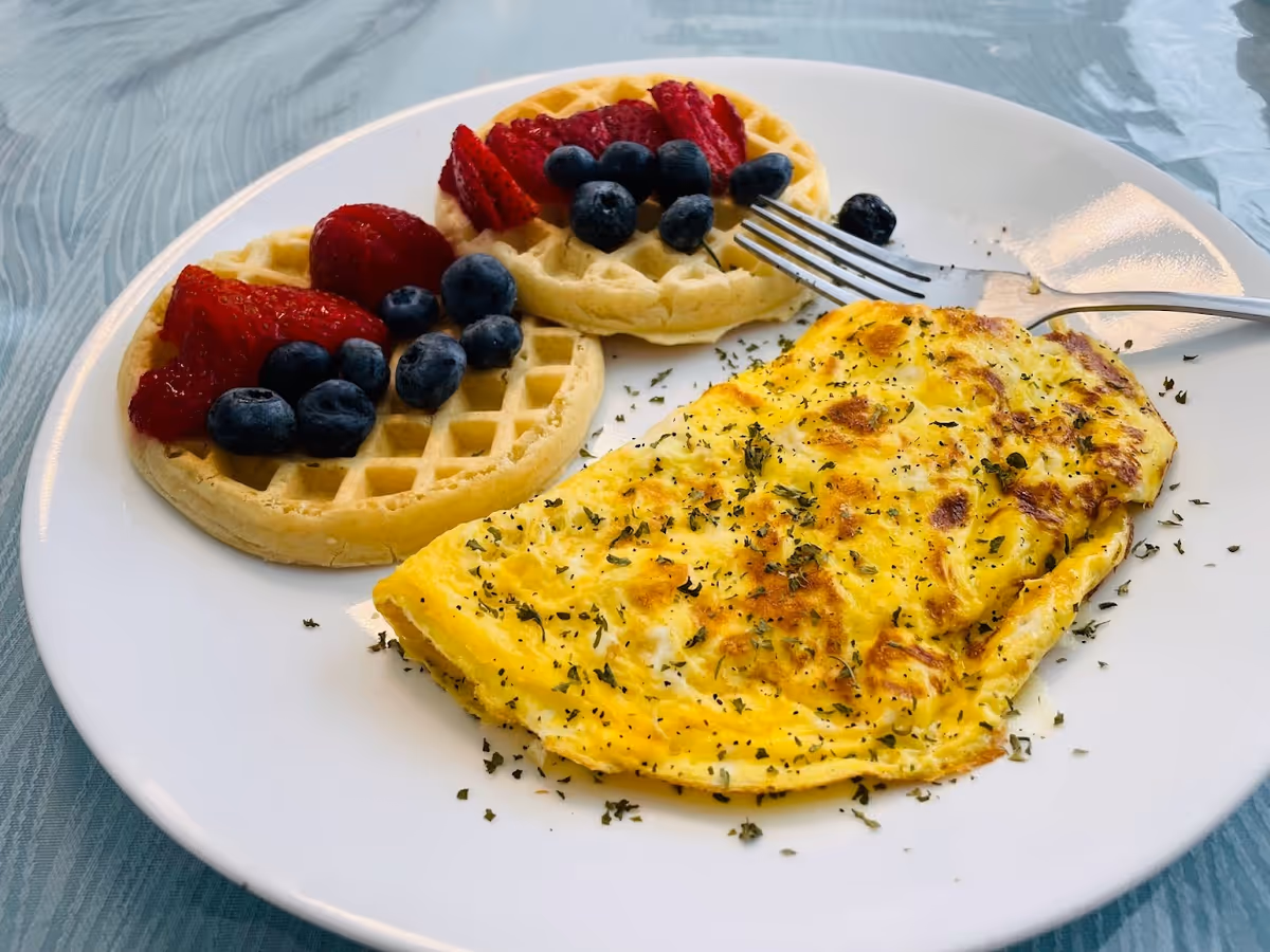 White plate with an omelet and two mini waffles topped with strawberries and blueberries on a blue tablecloth.