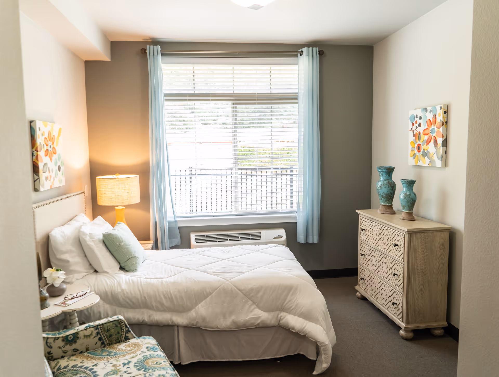 A cozy bedroom with a neatly made bed featuring white bedding and pillows. There is a bedside table with a lamp and a small flower vase. A patterned armchair is placed near the bed. The room has a large window with light blue curtains, allowing natural light to brighten the space. Across from the bed is a wooden dresser with two decorative blue vases and a colorful floral painting on the wall above it.
