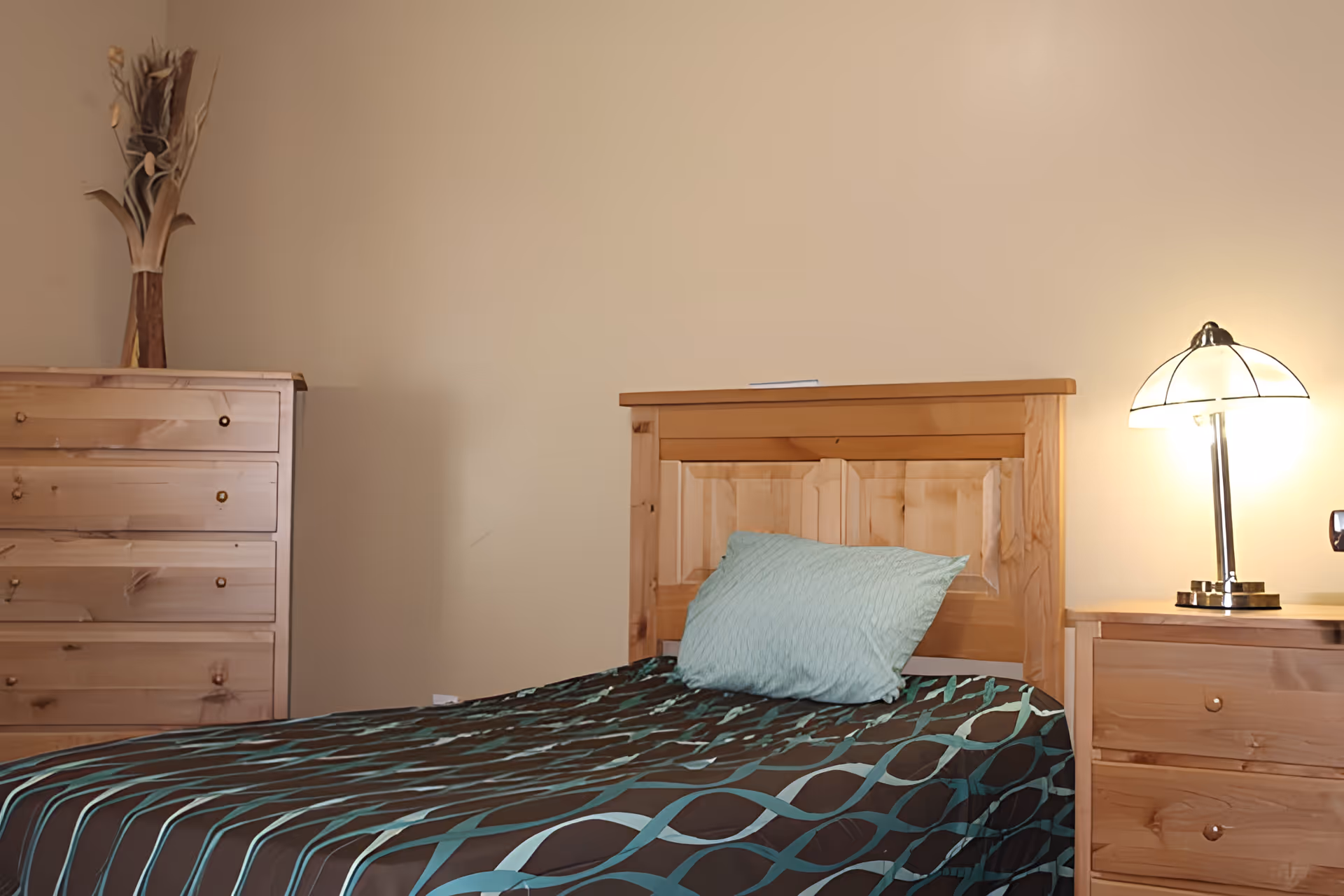 A modest bedroom featuring a wooden headboard and matching dresser, a single bed with teal-accented bedding, and a lit bedside lamp.