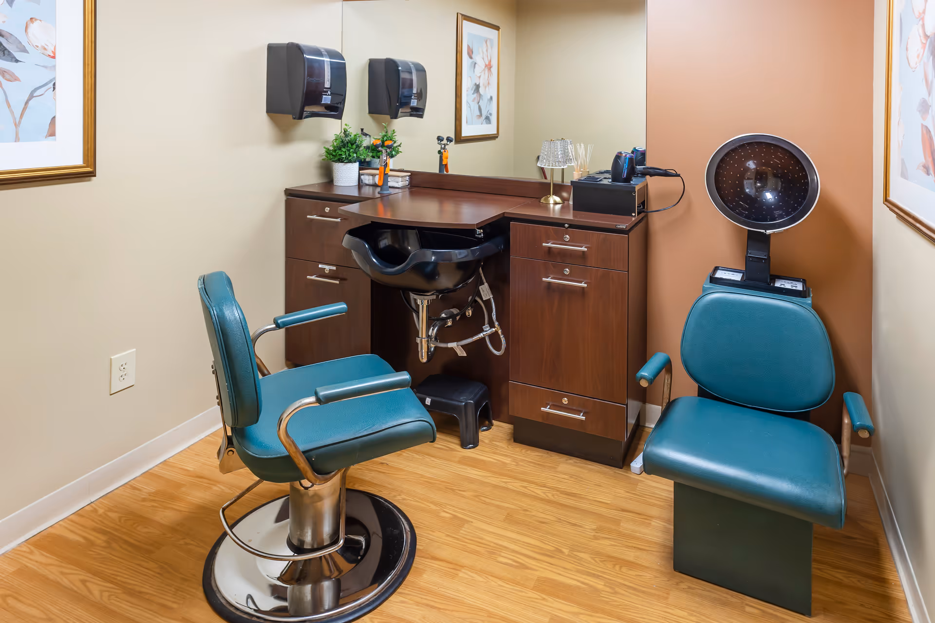 A small salon area with two teal salon chairs, a black hair washing sink, wooden cabinets with drawers, a hair dryer, and a hooded hair dryer. The walls are beige with framed floral artwork, and the floor is wood.