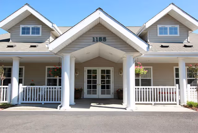 Front entrance of a light-gray senior living building with a covered porch, white columns, double glass doors and the number 1155 above the gable.
