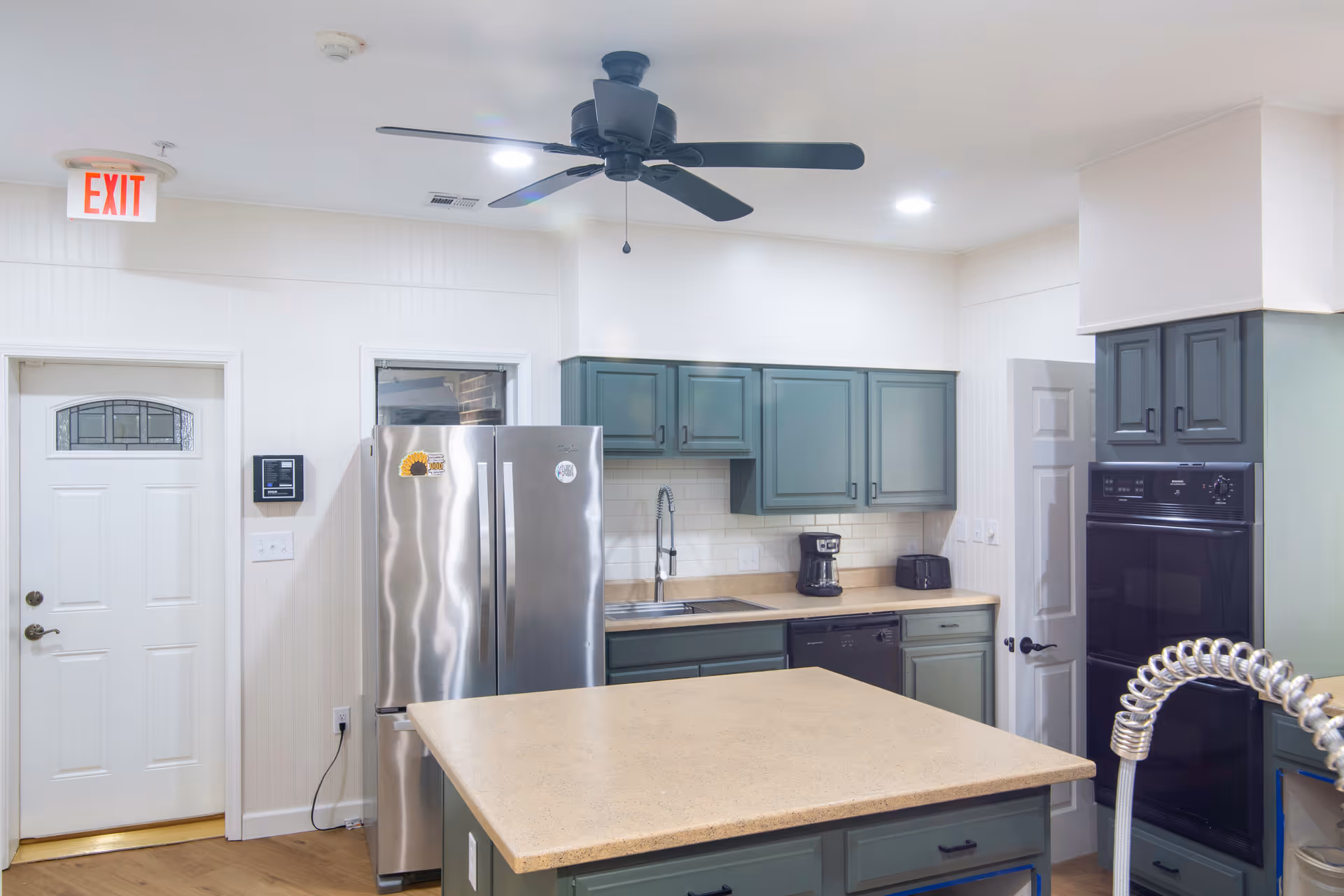 A modern kitchen with green cabinets, a large central island with a beige countertop, stainless steel refrigerator, built-in oven, black dishwasher, coffee maker, and toaster. The kitchen has white walls, a ceiling fan, and an exit door with a window.