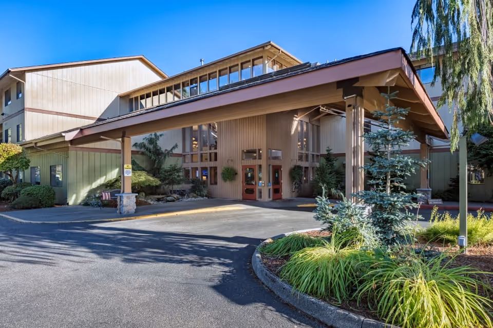 Exterior view of The Firs senior living facility entrance with a covered drop-off area, surrounded by greenery and trees under a clear blue sky.