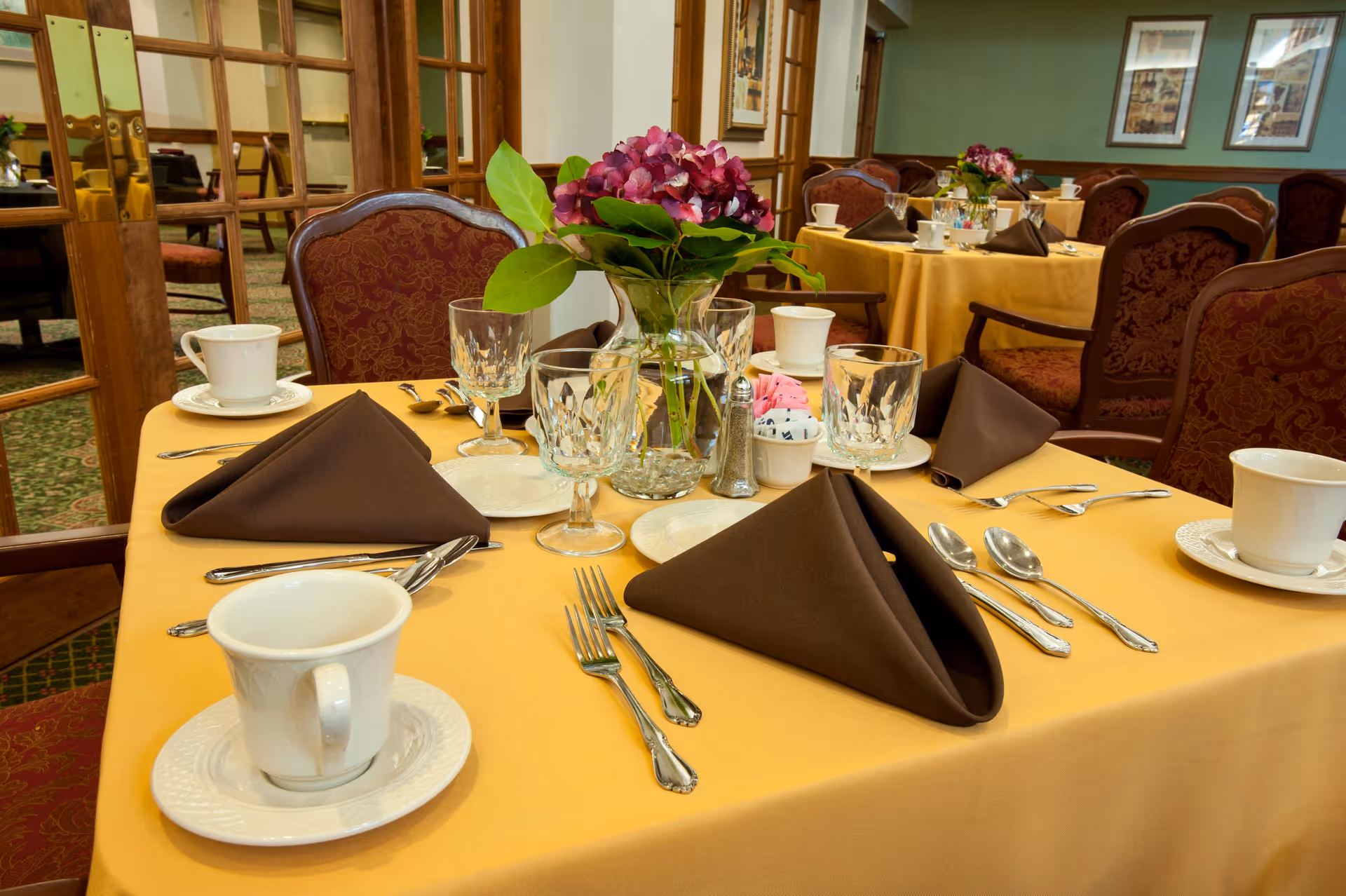 A dining room table set with a yellow tablecloth, folded brown napkins, crystal glasses, white cups, and a vase of purple flowers.