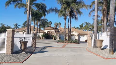 Wide driveway entrance leading to a large single-story building with a tiled roof, surrounded by tall palm trees and white fencing on both sides under a clear blue sky.