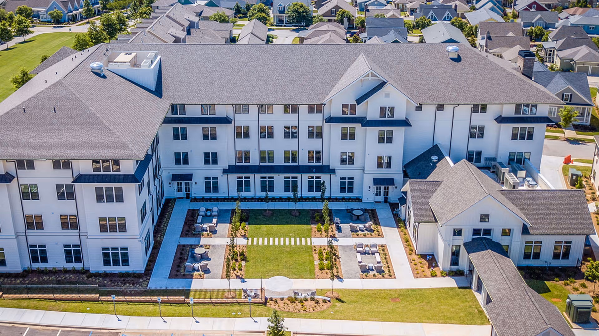 Aerial view of a large, modern senior living facility named Everlan of Clemson. The building is white with a gray roof and multiple windows. There is a landscaped courtyard in the center with walkways, seating areas, and greenery. Surrounding the facility are residential houses and green spaces.