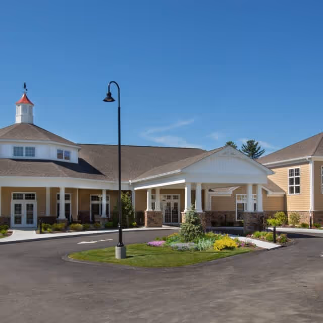 Exterior view of a single-story assisted living facility building with a covered entrance, landscaped garden with flowers and shrubs, a roundabout driveway, and a clear blue sky.