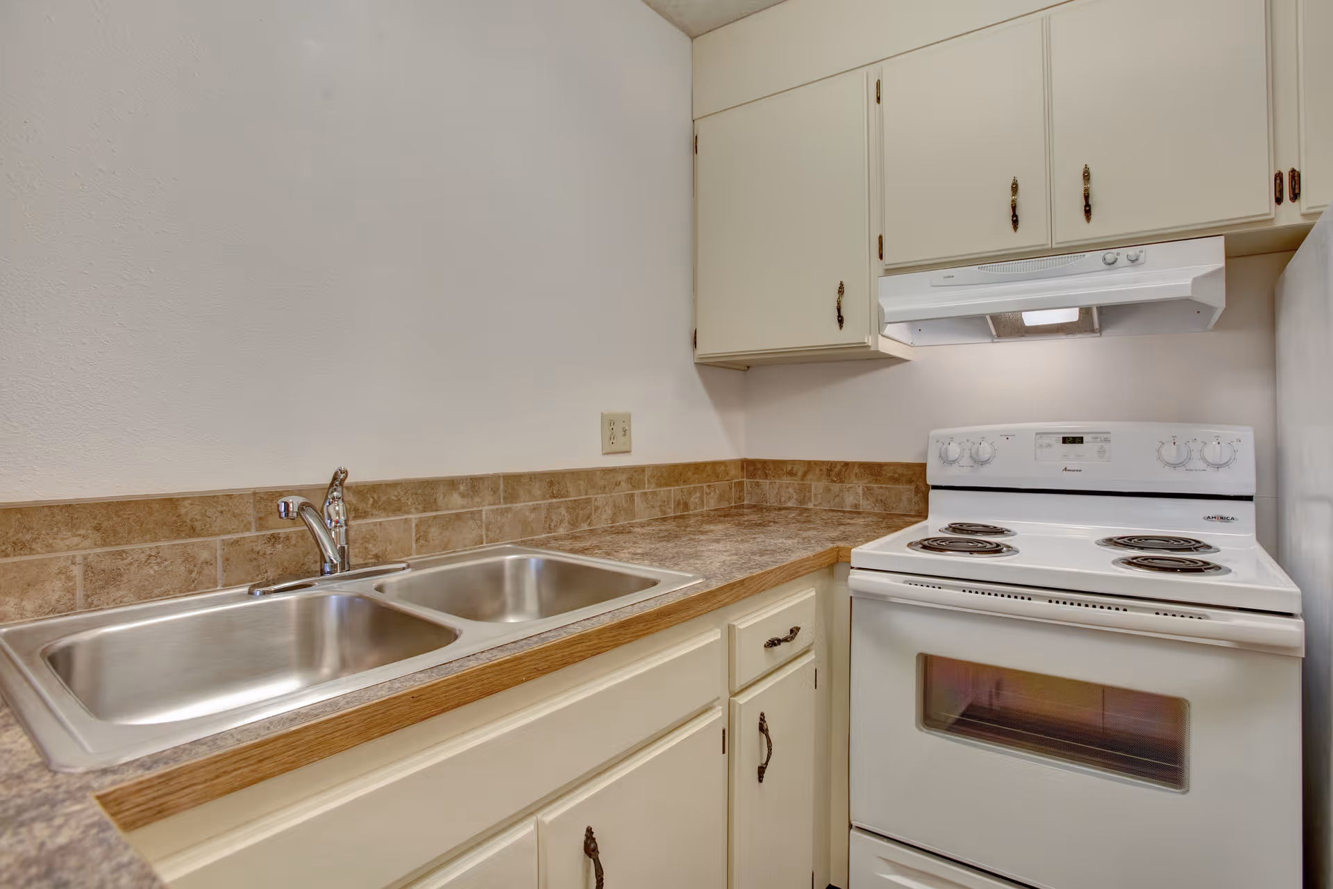 Small kitchen with a double stainless steel sink, laminate countertops, cream cabinets, and a white electric stove with range hood.
