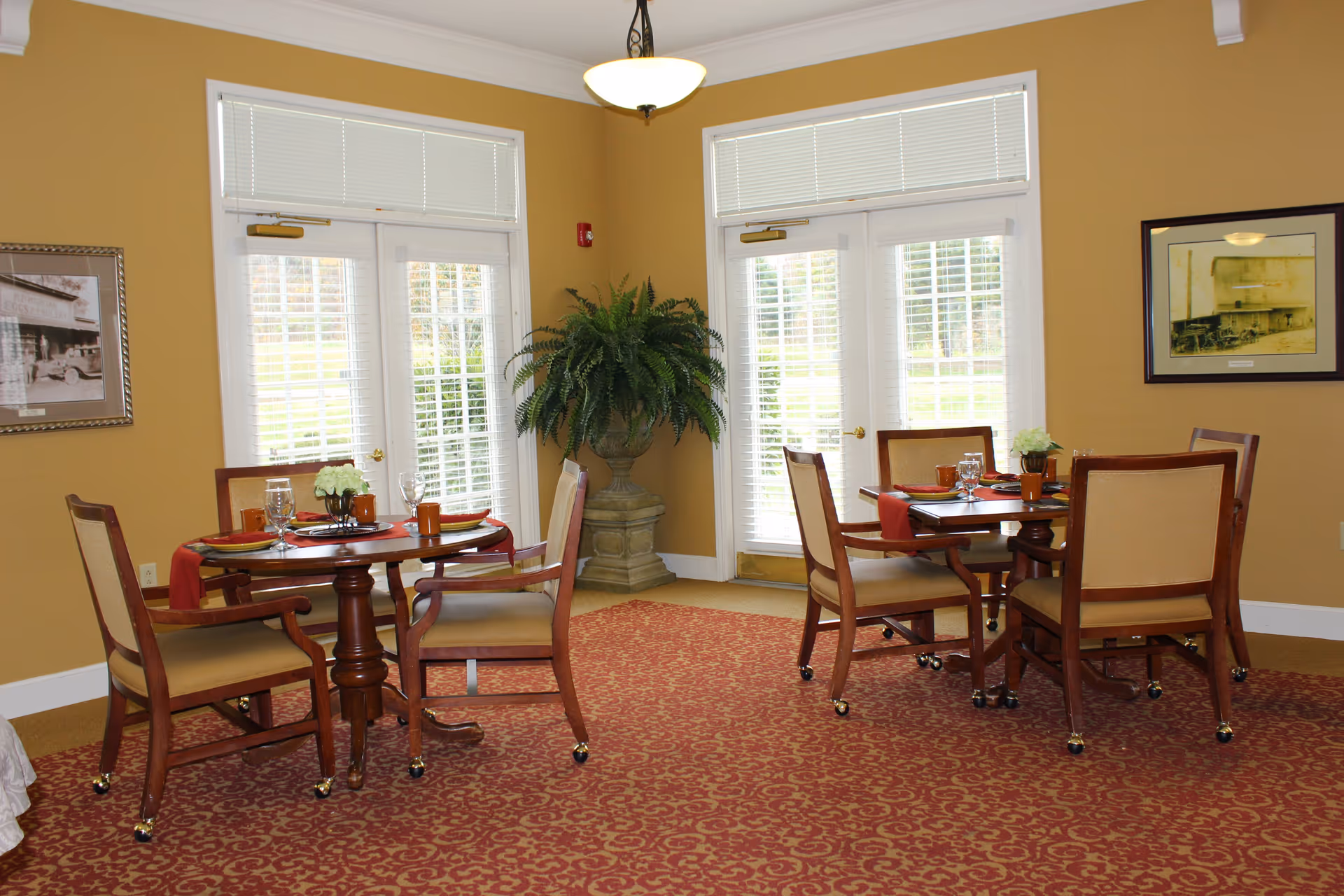 Dining room with two round tables set for meals, wooden upholstered chairs, large windows with blinds, and a potted fern.