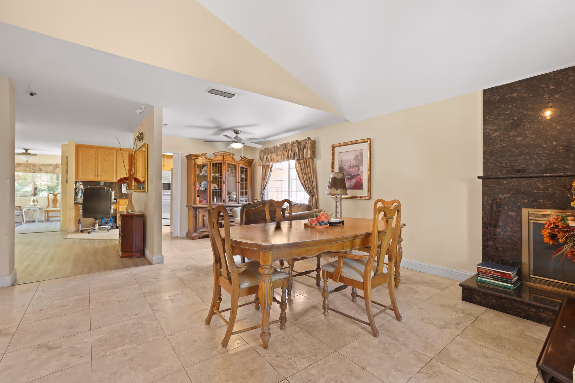 Wooden dining table with four chairs in a bright dining area adjoining a living space and kitchen, with a china cabinet and fireplace visible.