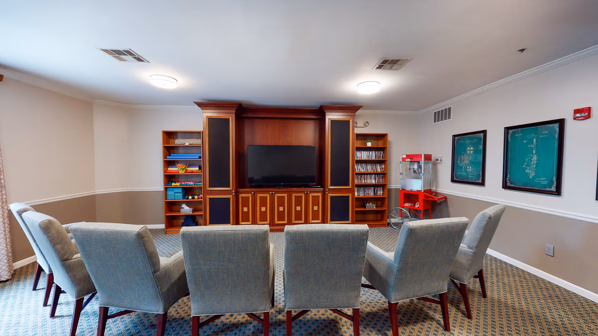 A cozy common room with a row of seven upholstered chairs facing a large wooden entertainment center with a flat-screen TV. The entertainment center has shelves with DVDs and toys. On the right side, there is a red popcorn machine and framed artwork on the wall. The room has patterned carpet and neutral-colored walls with white trim.