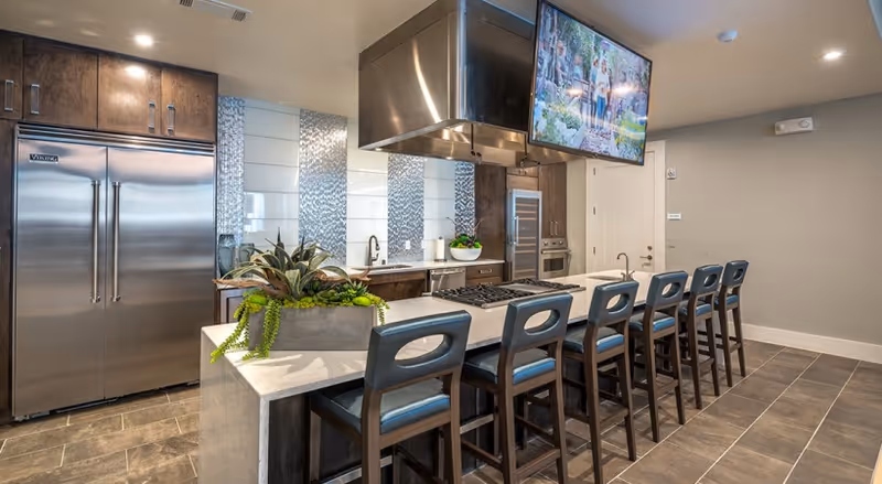 Modern kitchen with a long island featuring six high chairs, a built-in stove, and a large stainless steel refrigerator. The kitchen has dark wooden cabinets, a stainless steel range hood, and a mounted flat-screen TV. Decorative plants are placed on the island, and the floor is tiled.