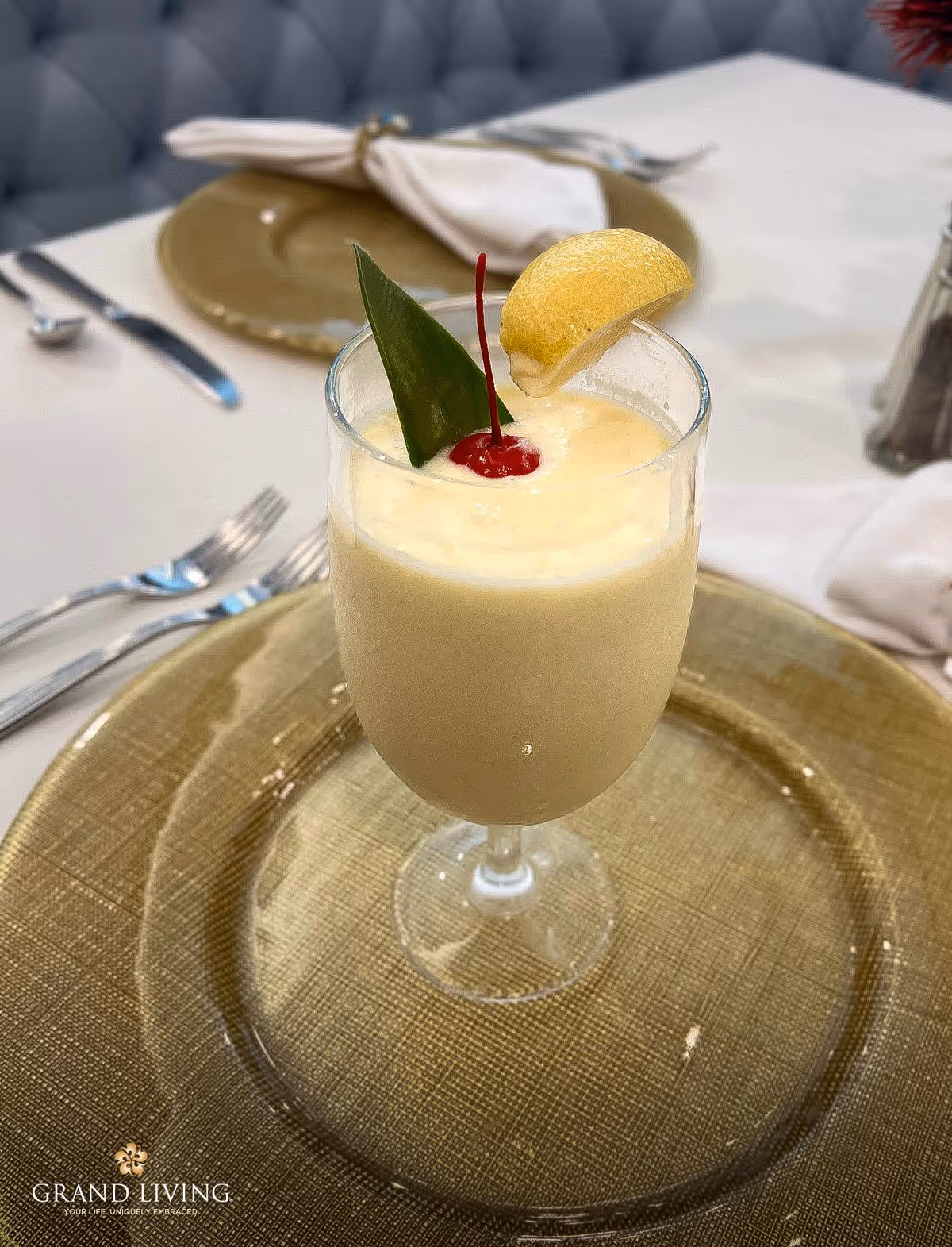 A creamy garnished cocktail with a lemon slice and cherry on a gold charger plate at a set dining table.