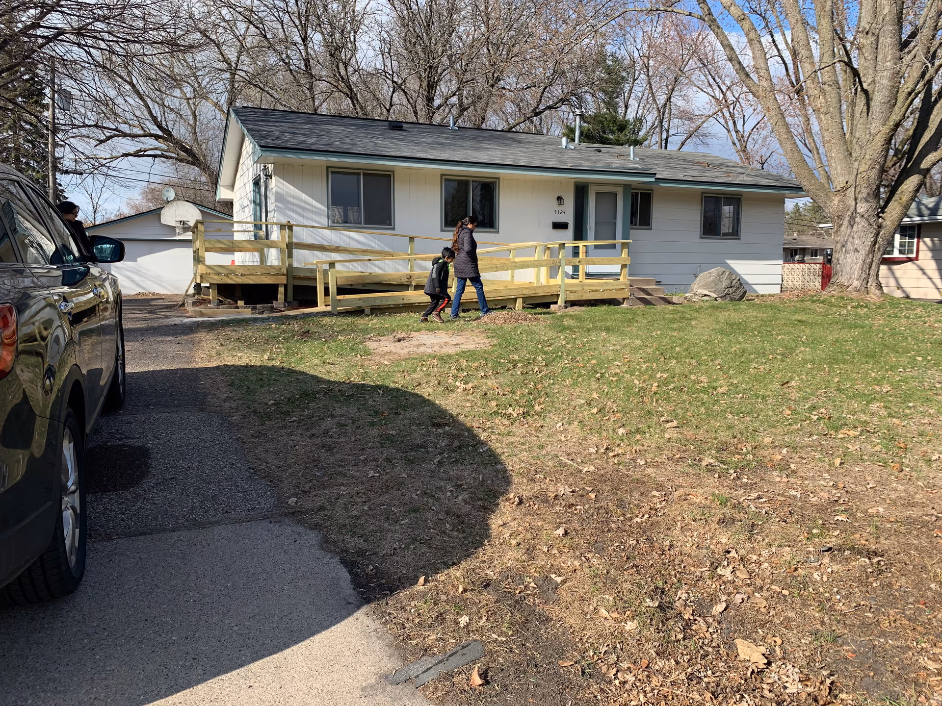 A single-story white house with a gray roof and a wooden wheelchair ramp leading to the front door. A woman and a child are walking on the grass in front of the house. There is a black car parked on the driveway to the left, and leafless trees are visible in the background.