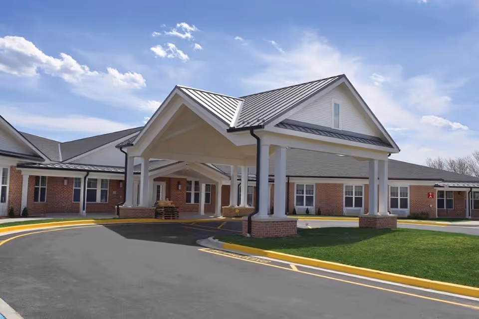Front entrance of a brick senior living facility with a covered porte-cochere, driveway, and manicured lawn.