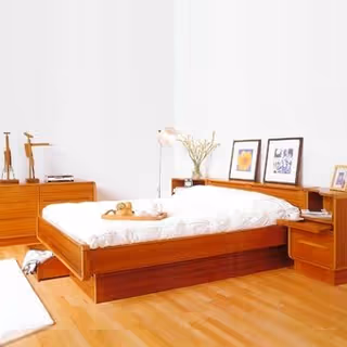 A minimalist bedroom with a wooden bed frame and white bedding. The room features wooden furniture including a dresser and nightstand, with framed pictures and a vase with flowers on the headboard shelf. The floor is light wood, and the walls are plain white.