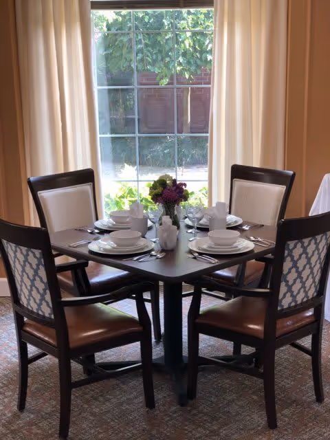 A dining table set for four with white plates, bowls, cups, and silverware. A small flower arrangement is placed in the center of the dark wood table. Four chairs with patterned backs and brown seats surround the table. Behind the table is a large window with beige curtains, showing greenery outside.