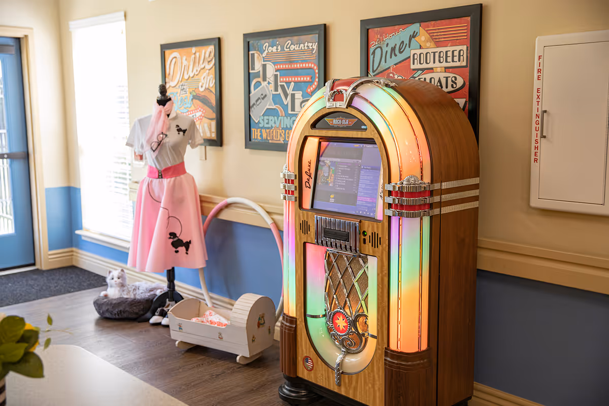 A vintage-style jukebox with colorful lights stands against a wall in a room decorated with retro diner-themed posters. To the left, there is a mannequin dressed in a 1950s-style pink skirt and white top with a poodle design, and a small cradle with a doll inside. A cat bed with a white cat is on the floor near a window with blinds. The room has light-colored walls with a blue lower half and a fire extinguisher cabinet on the right side.