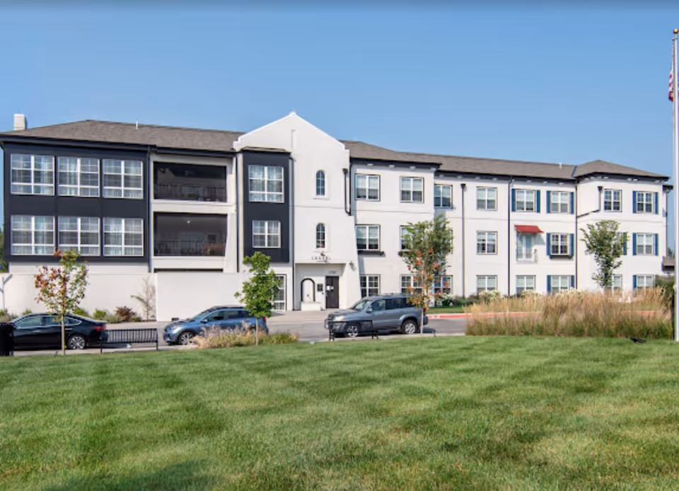 Three-story senior living building with a green lawn and parked cars in front under a clear blue sky.