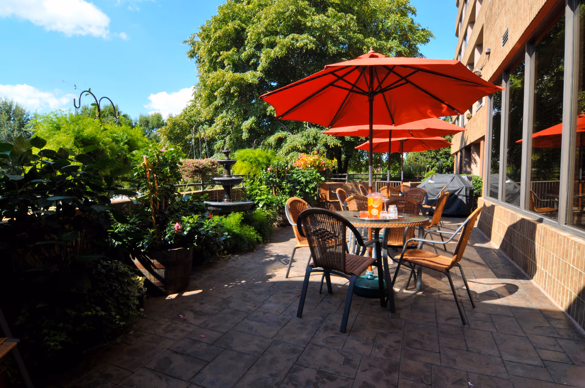 Outdoor patio area with several round tables and wicker chairs under large red umbrellas. The patio is surrounded by lush green plants and trees, with a multi-tiered water fountain visible in the background. The building with large windows is on the right side, and the sky is clear and blue.