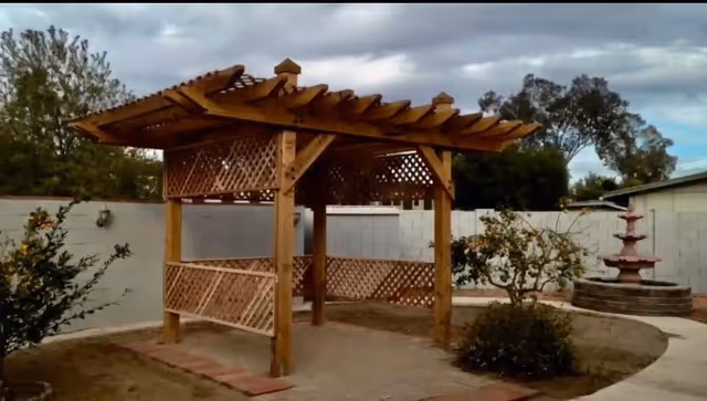 Outdoor area featuring a wooden pergola with lattice sides, surrounded by small bushes and plants, a curved paved walkway, and a multi-tiered water fountain in the background under a cloudy sky.