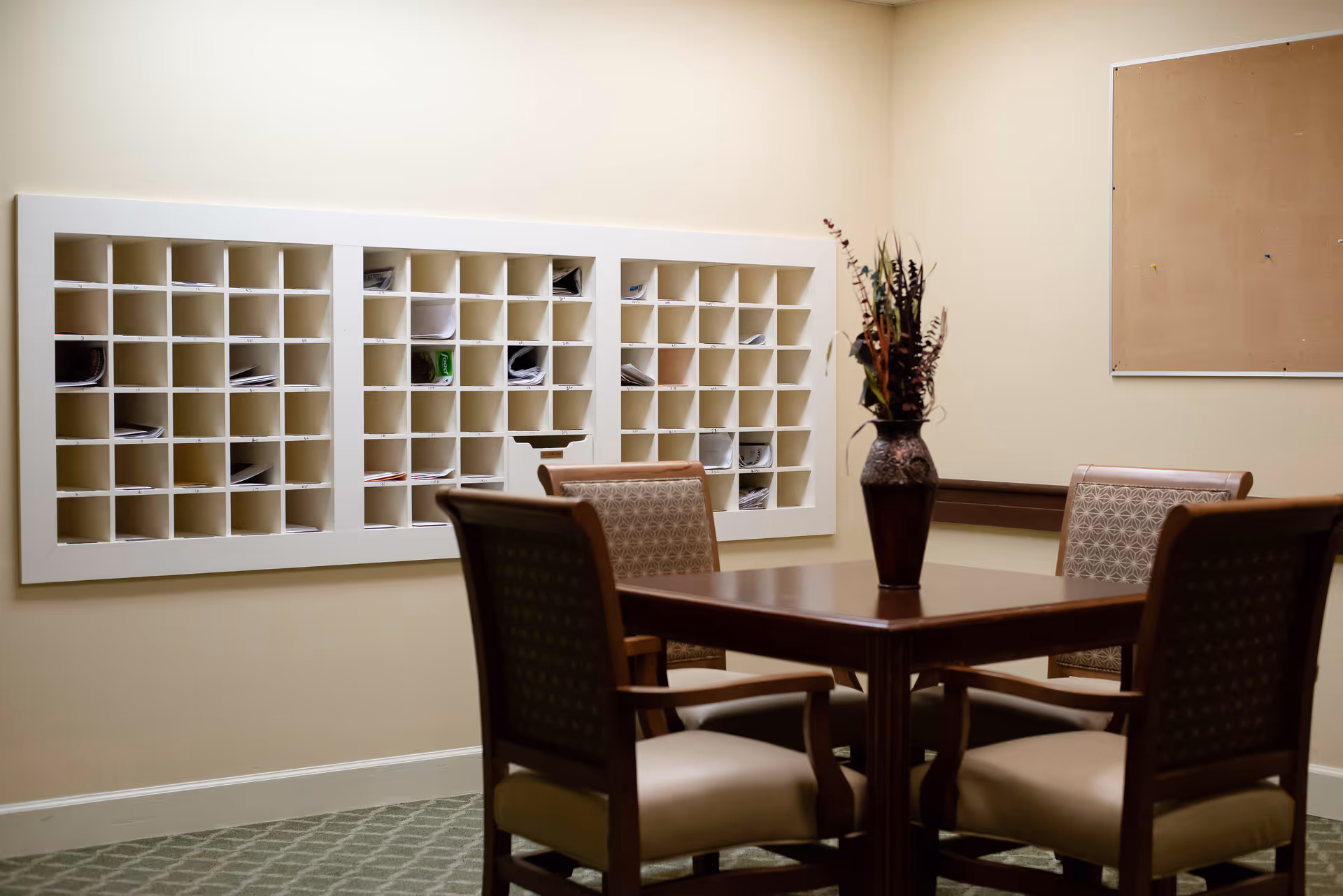 A small room with a wooden table and four cushioned chairs arranged around it. On the wall behind the table is a large white mail organizer with multiple slots containing papers. There is also a decorative vase with dried flowers on the table and a cork bulletin board on the wall.