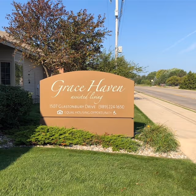 Outdoor view of a large brown sign for Grace Haven assisted living, located at 1507 Glastonbury Drive with phone number (989) 224-1650. The sign also displays the Equal Housing Opportunity logo and is surrounded by green grass, bushes, and trees, with a sidewalk and road visible to the right.