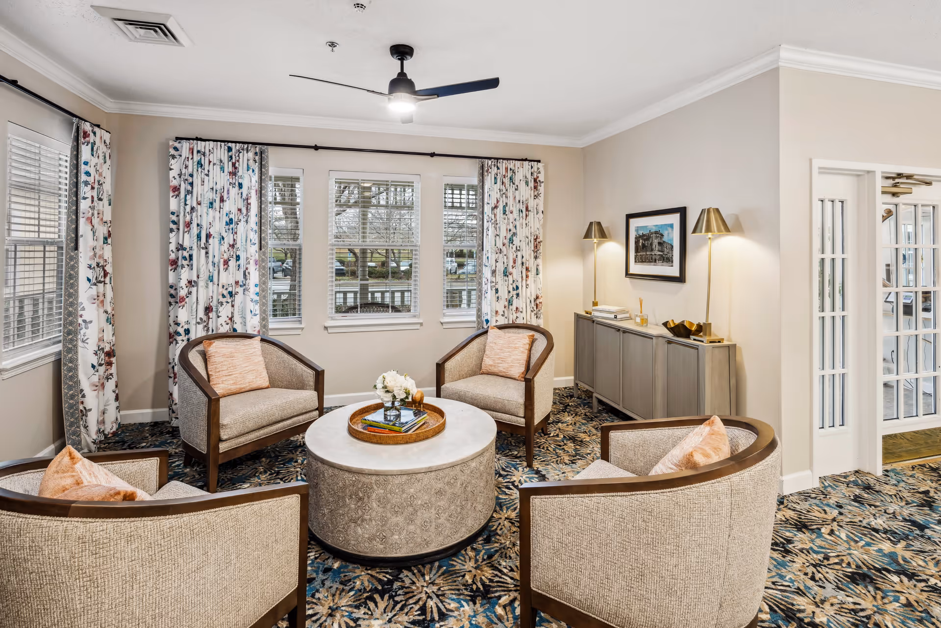 A cozy sitting area with four beige armchairs arranged around a round coffee table with a decorative tray and flowers. The room has floral patterned curtains on windows, a patterned carpet, a sideboard with two gold lamps, and a framed picture on the wall. There is a ceiling fan with a light fixture above.