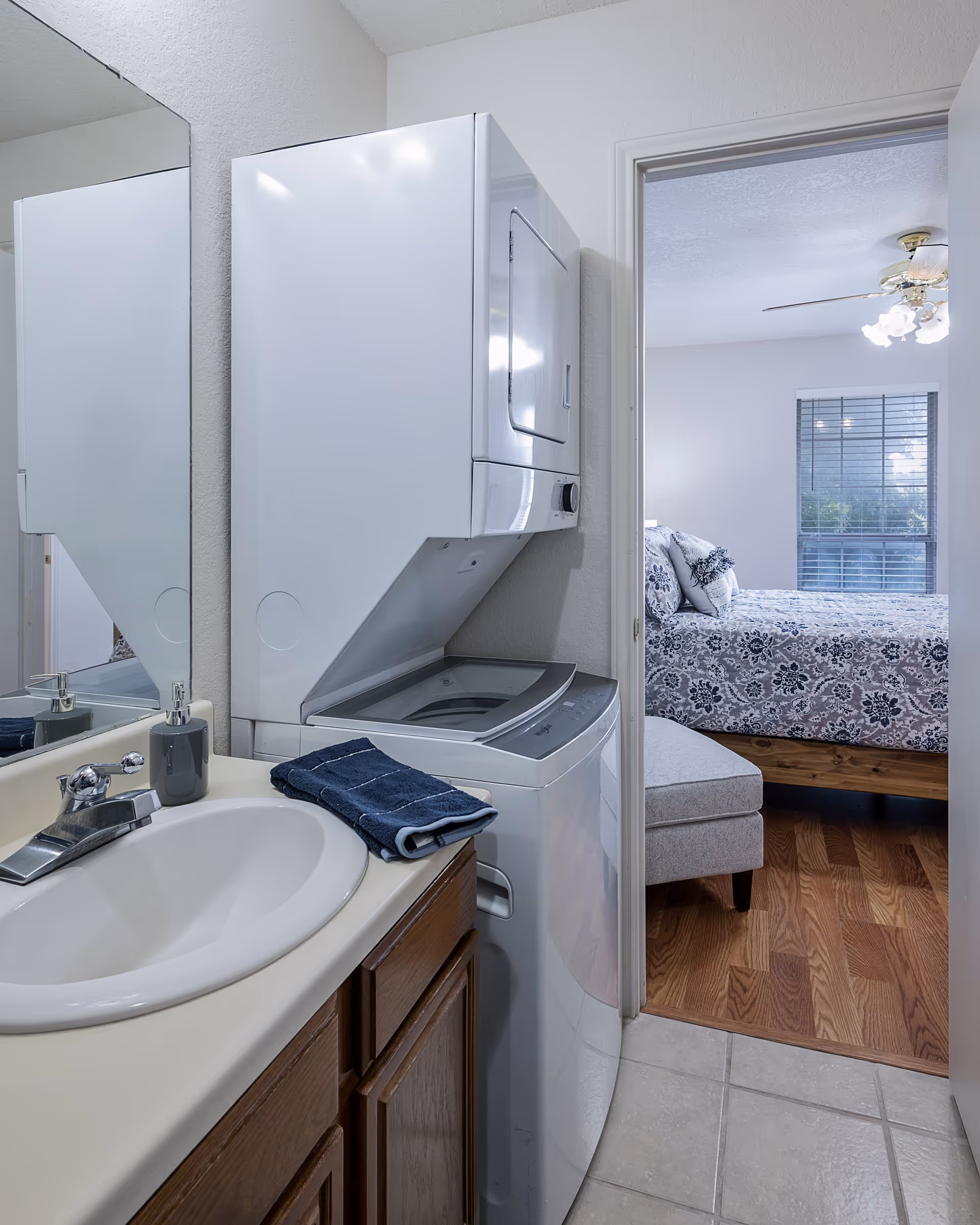 A small laundry area with a stacked washer and dryer next to a bathroom sink with a faucet and a dark blue towel. Through an open door, a bedroom with a bed covered in a floral patterned bedspread and a window with blinds is visible.