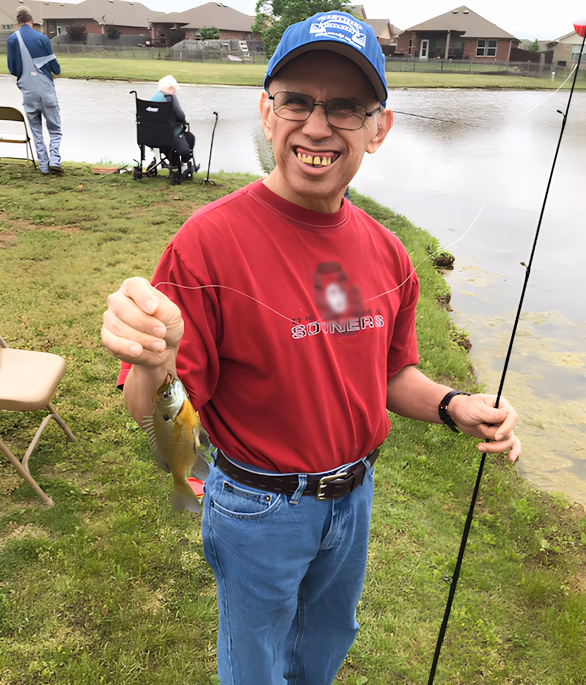 A man wearing a blue cap, glasses, a red shirt, and blue jeans is smiling and holding a fishing rod in one hand and a small fish in the other. He is standing on grassy ground near a pond. In the background, there is a person in a wheelchair facing the pond and another person standing nearby. Houses are visible across the pond.