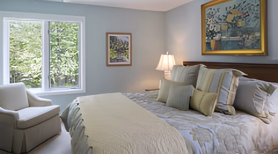 A cozy bedroom with a large bed covered in a beige and light green bedspread and multiple pillows. There is a white armchair near a window with a view of green trees outside. The walls are painted light blue and decorated with two framed paintings. A bedside table with a lamp is also visible.