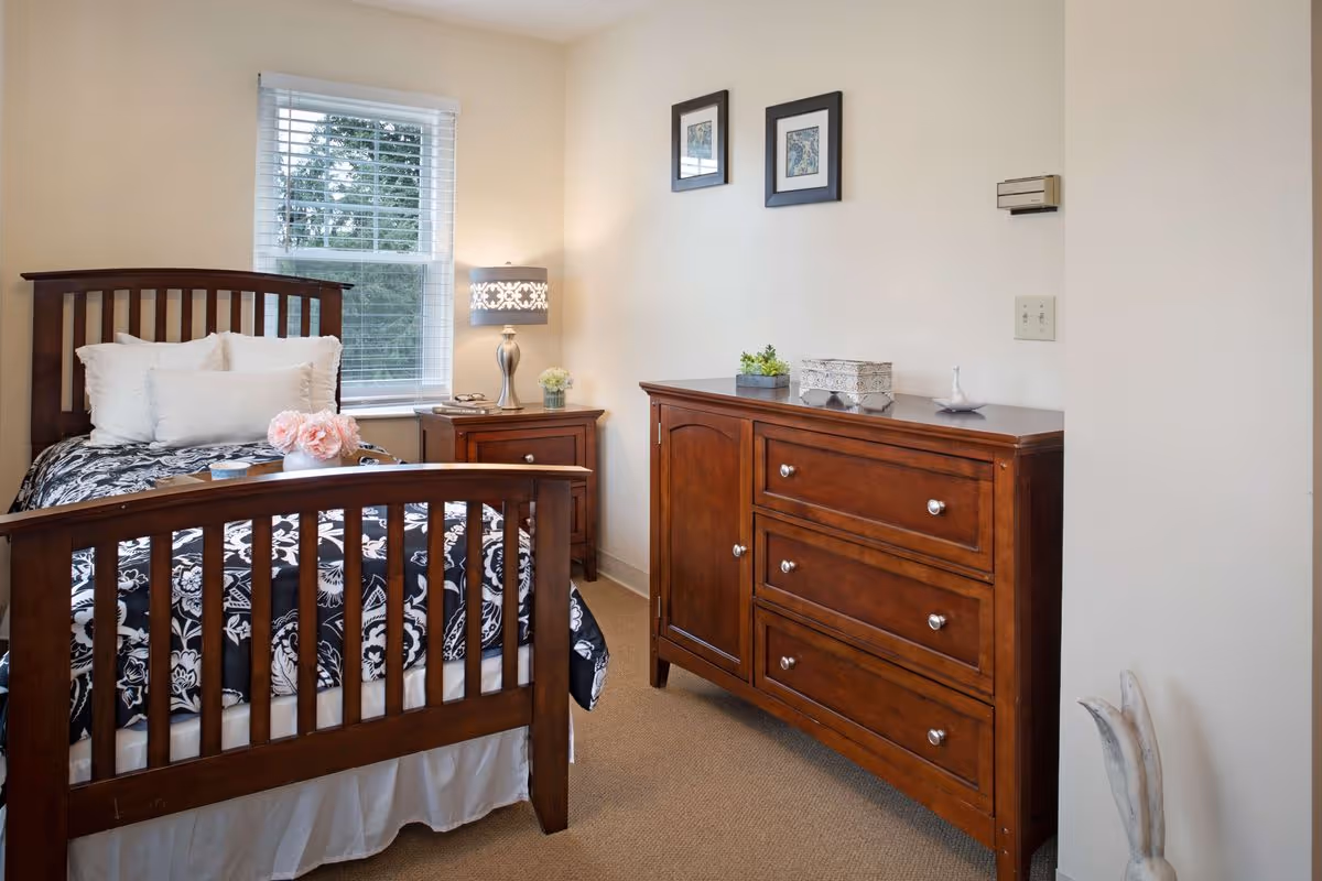 A cozy bedroom with a wooden bed dressed in black-and-white patterned bedding, a nightstand and lamp by a window, and a wooden dresser against the wall.