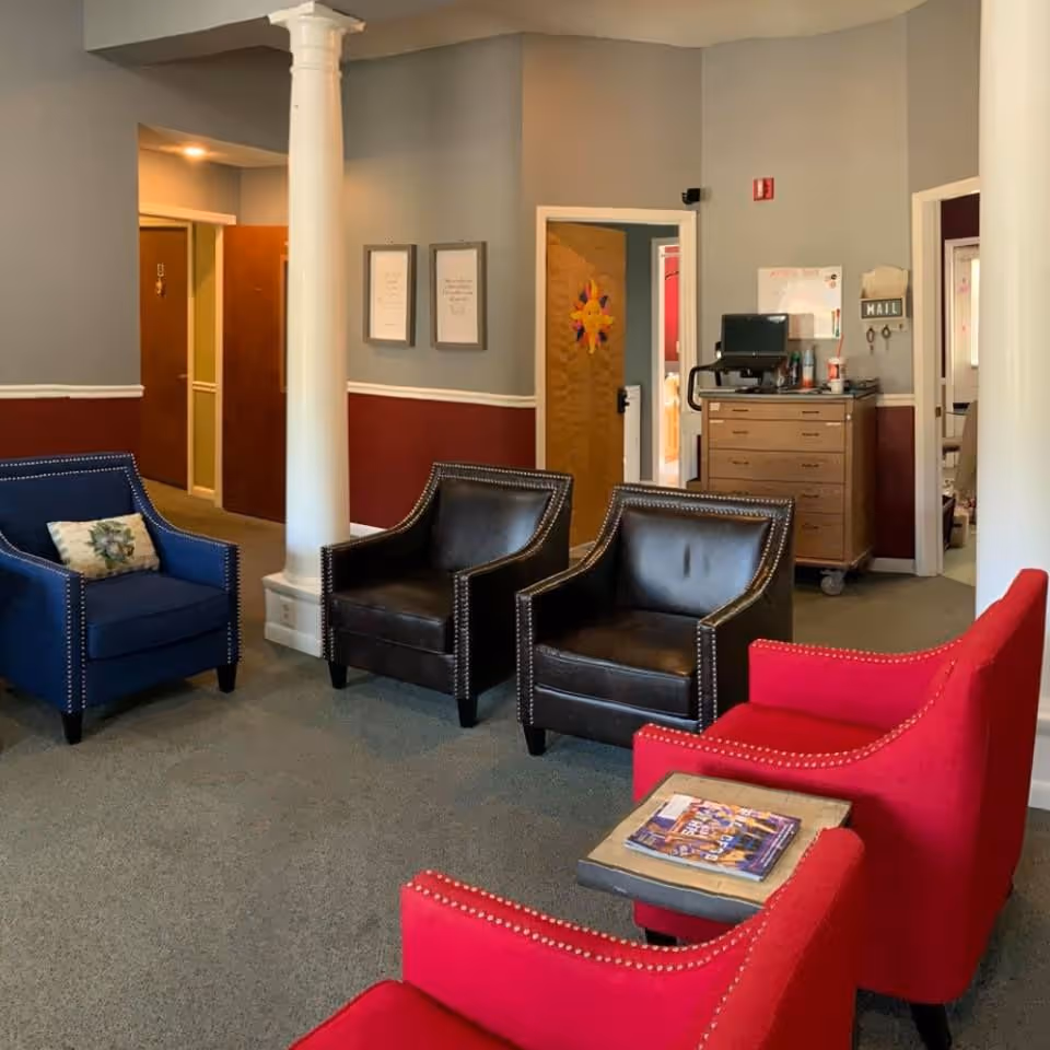 A cozy sitting area in an assisted living facility with two red armchairs, two black leather armchairs, and one blue armchair arranged around a small table with magazines. The room has gray and maroon walls, white columns, and several doorways leading to other rooms.