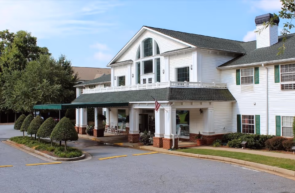 Exterior view of a white two-story building with green shutters and a green roof, featuring a covered entrance with columns, rocking chairs, and an American flag. The building is surrounded by neatly trimmed bushes and trees under a clear blue sky.