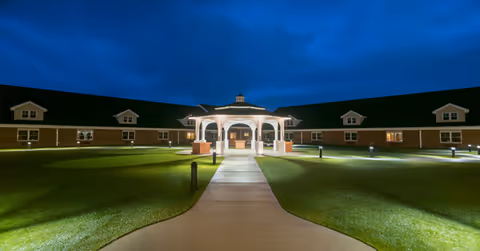 Nighttime view of Armstrong Memory Care Assisted Living facility showing a well-lit gazebo in the center with a paved walkway leading to it, surrounded by a large grassy courtyard and a single-story building with multiple windows in the background under a dark blue sky.