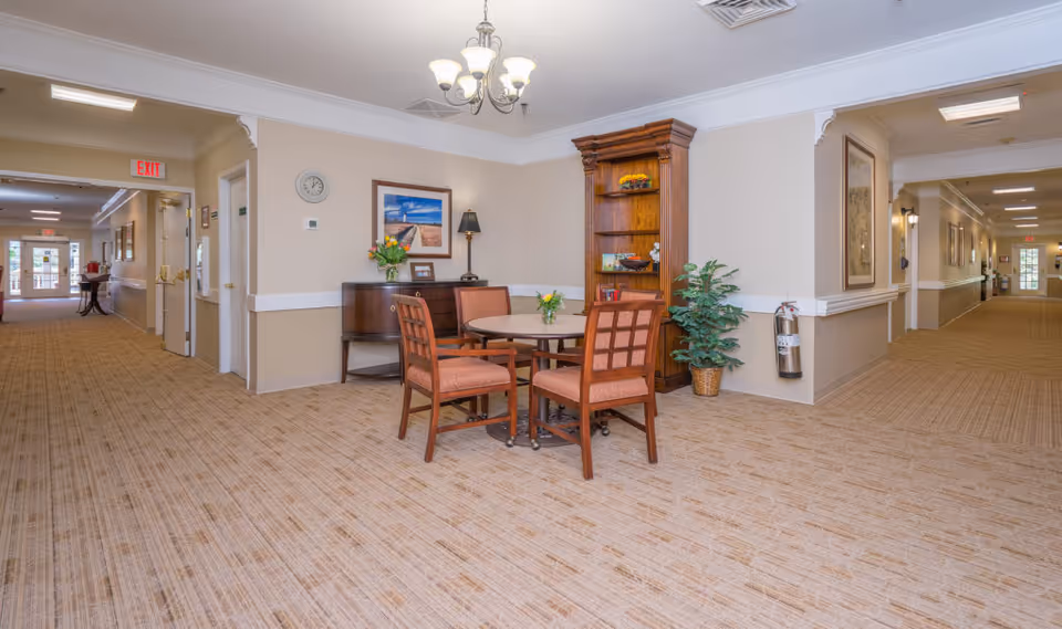 A spacious hallway in a senior living facility with beige walls and carpeted floors. In the center, there is a round table with four wooden chairs upholstered in orange fabric. Behind the table is a wooden bookshelf with decorative items and a small sideboard with a lamp and a vase of flowers. The hallway extends in both directions with exit signs and doors visible.