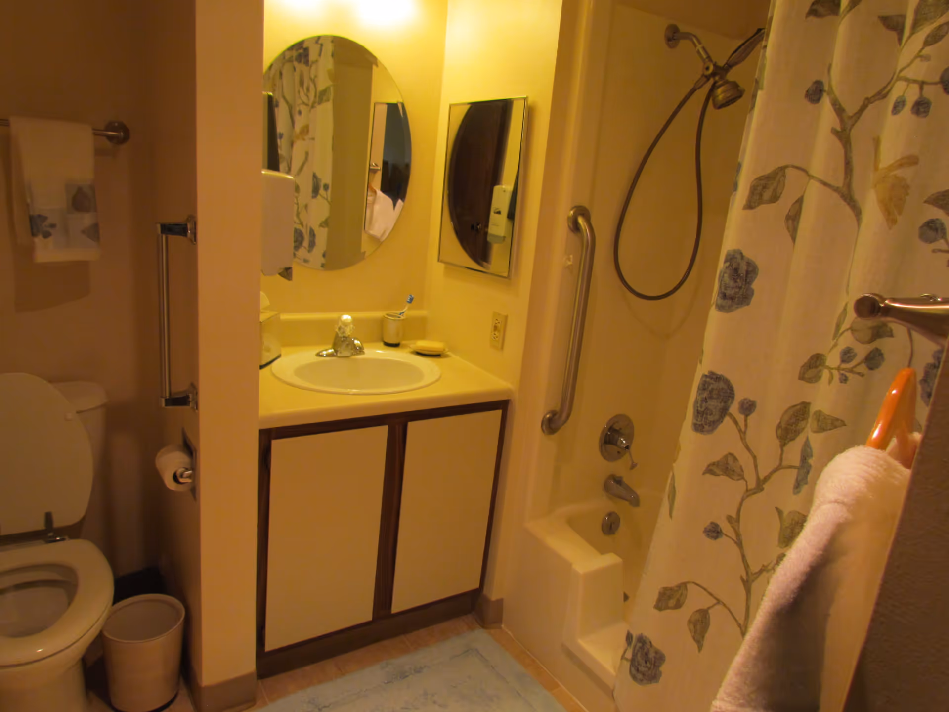 A bathroom with a toilet, a sink with a cabinet underneath, a round mirror above the sink, a shower with a floral-patterned curtain, and grab bars for accessibility. There is a towel hanging on a rack and a small trash bin next to the toilet.
