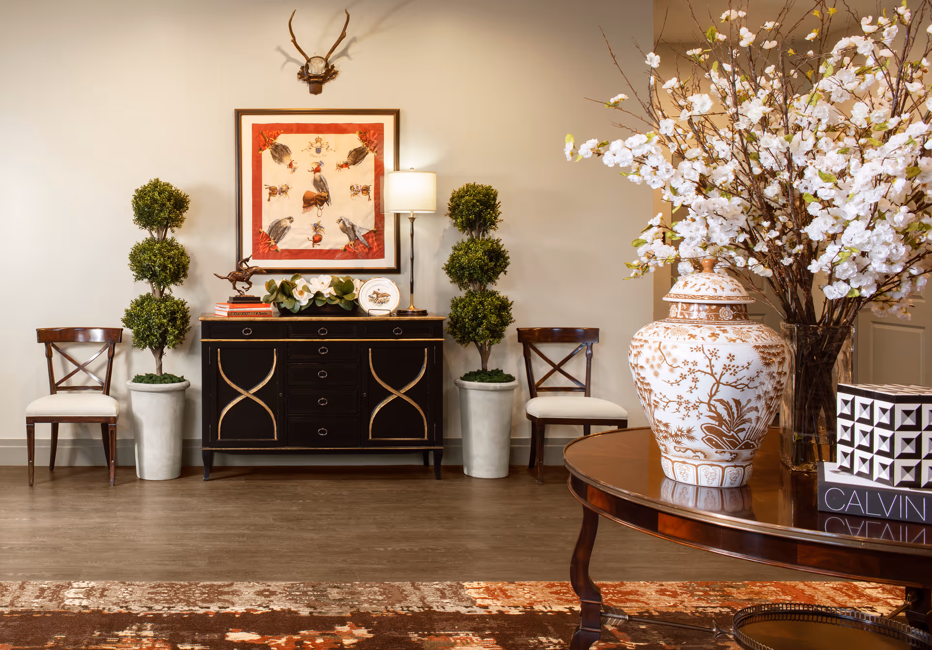 A stylish interior room featuring a black sideboard with gold accents, flanked by two potted topiary plants and two wooden chairs with white cushions. Above the sideboard hangs a framed artwork depicting birds and insects, with a mounted antler decoration above it. To the right, a round wooden table holds a large decorative vase with white flowers and a black and white patterned box. The floor is covered with a patterned rug.