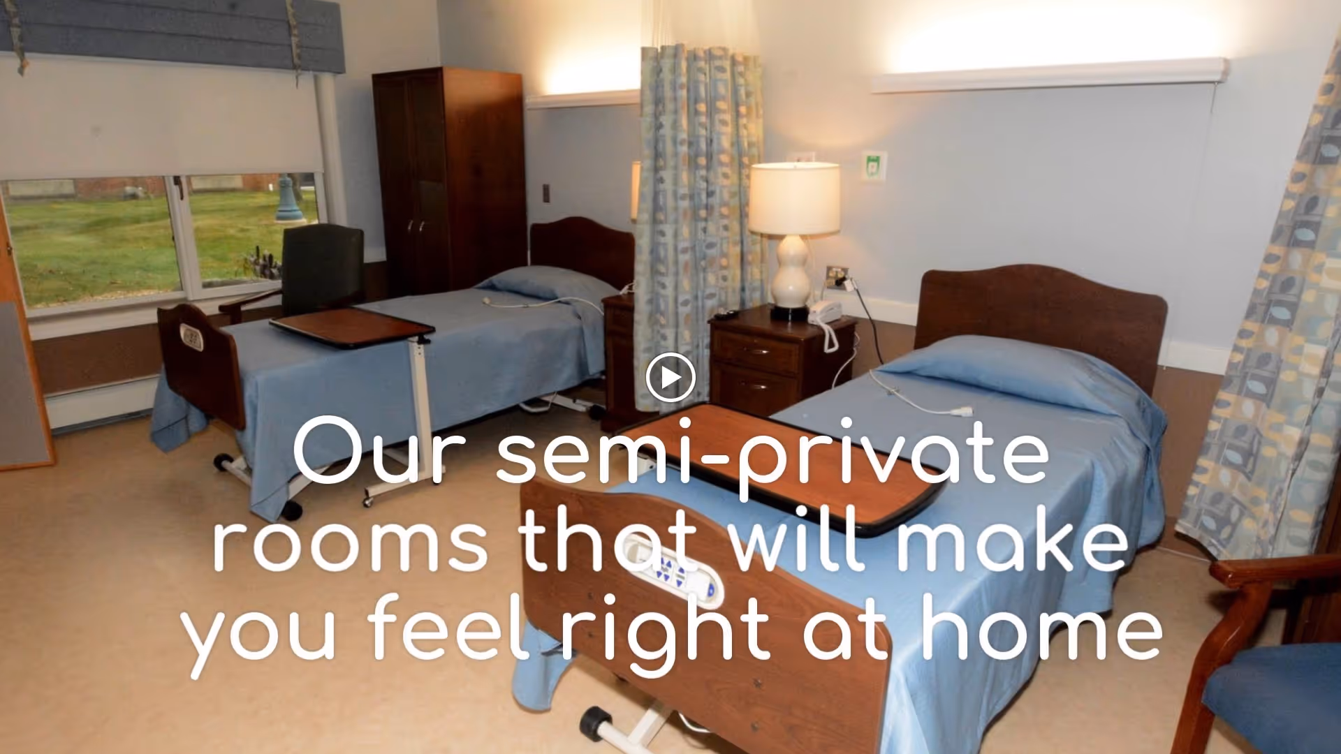 Semi-private patient room with two hospital-style beds, bedside tables, lamps, curtains, and a window.