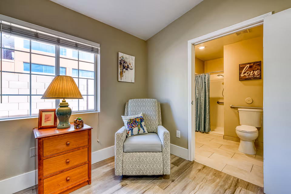 A cozy corner of a senior living facility room featuring a patterned armchair with a colorful elephant pillow, a wooden side table with a lamp, picture frame, and small decorative item, next to a window with blinds. An open doorway reveals a bathroom with a toilet, grab bars, a shower with a curtain, and a wall decoration that says 'love the little things.'