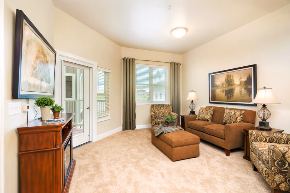 Cozy living room with a brown sofa, patterned armchairs, an ottoman, side tables with lamps, framed artwork, and a door to a balcony.