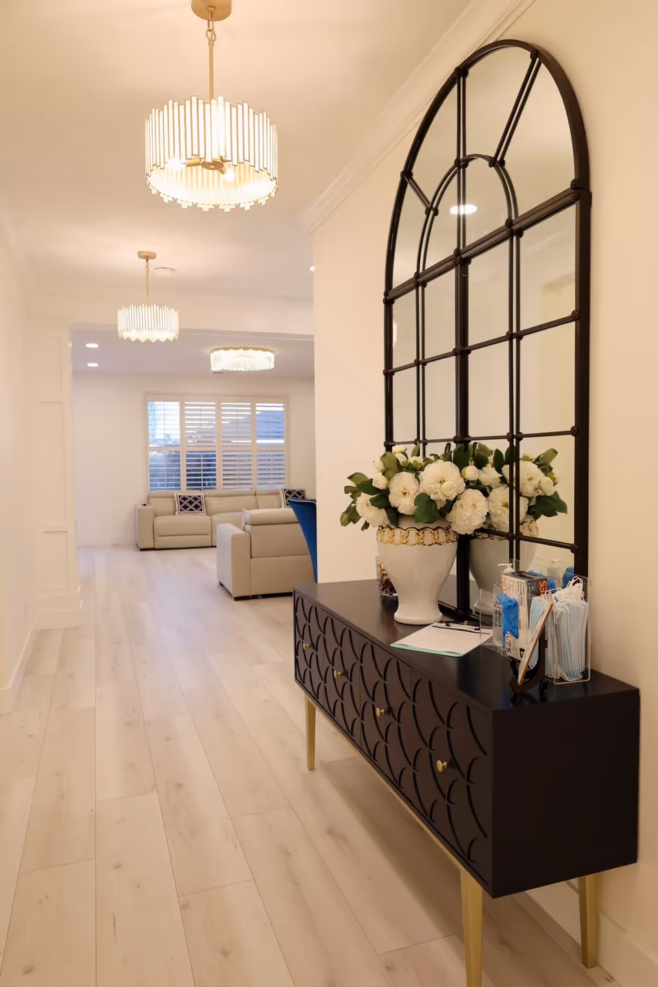 A bright and modern hallway with light wood flooring and white walls. On the right side, there is a black console table with gold legs, decorated with a white vase filled with white flowers, a large arched window-style mirror above it, and a container holding face masks and hand sanitizer. The hallway leads to a living room area with beige sofas and large windows with white shutters. Three elegant pendant lights hang from the ceiling.