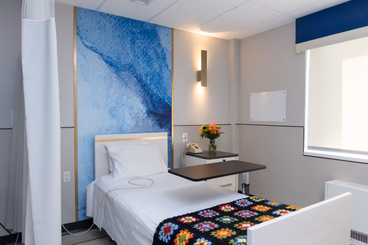 A clean and modern patient room with a single bed covered with white sheets and a colorful crocheted blanket at the foot. The bed is positioned against a wall with a large blue abstract artwork. Next to the bed is a small bedside table with a telephone and a vase of flowers. A wall-mounted light fixture provides soft lighting, and a window with a roller shade lets in natural light. A privacy curtain is partially visible on the left side of the image.
