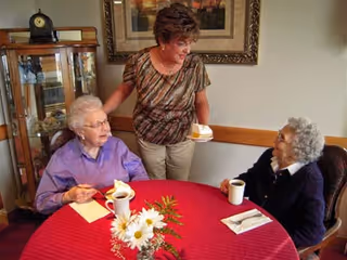 Three women seated around a round table with a red tablecloth, coffee cups, flowers, and a plate with dessert in an indoor dining area.