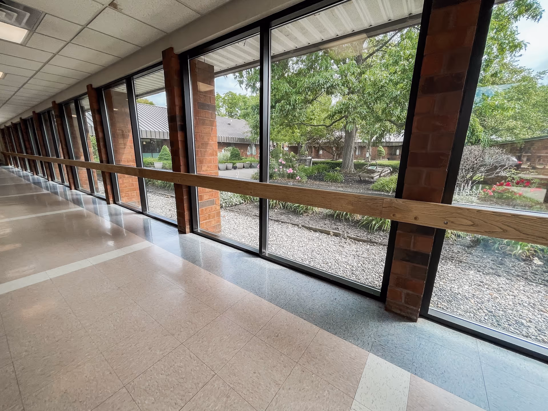 Long interior corridor with floor-to-ceiling windows and a wooden handrail overlooking a landscaped courtyard.