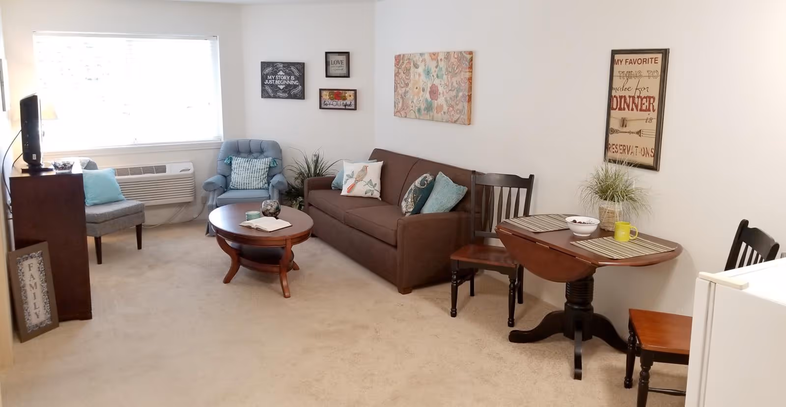A cozy living room with a brown sofa adorned with decorative pillows, a blue armchair, a wooden coffee table with an open book and decorative items, and a small dining table with two chairs set with placemats, a bowl, and a yellow mug. The room has beige carpet, white walls decorated with framed art and quotes, a window with blinds, and a TV on a stand next to a floor lamp.