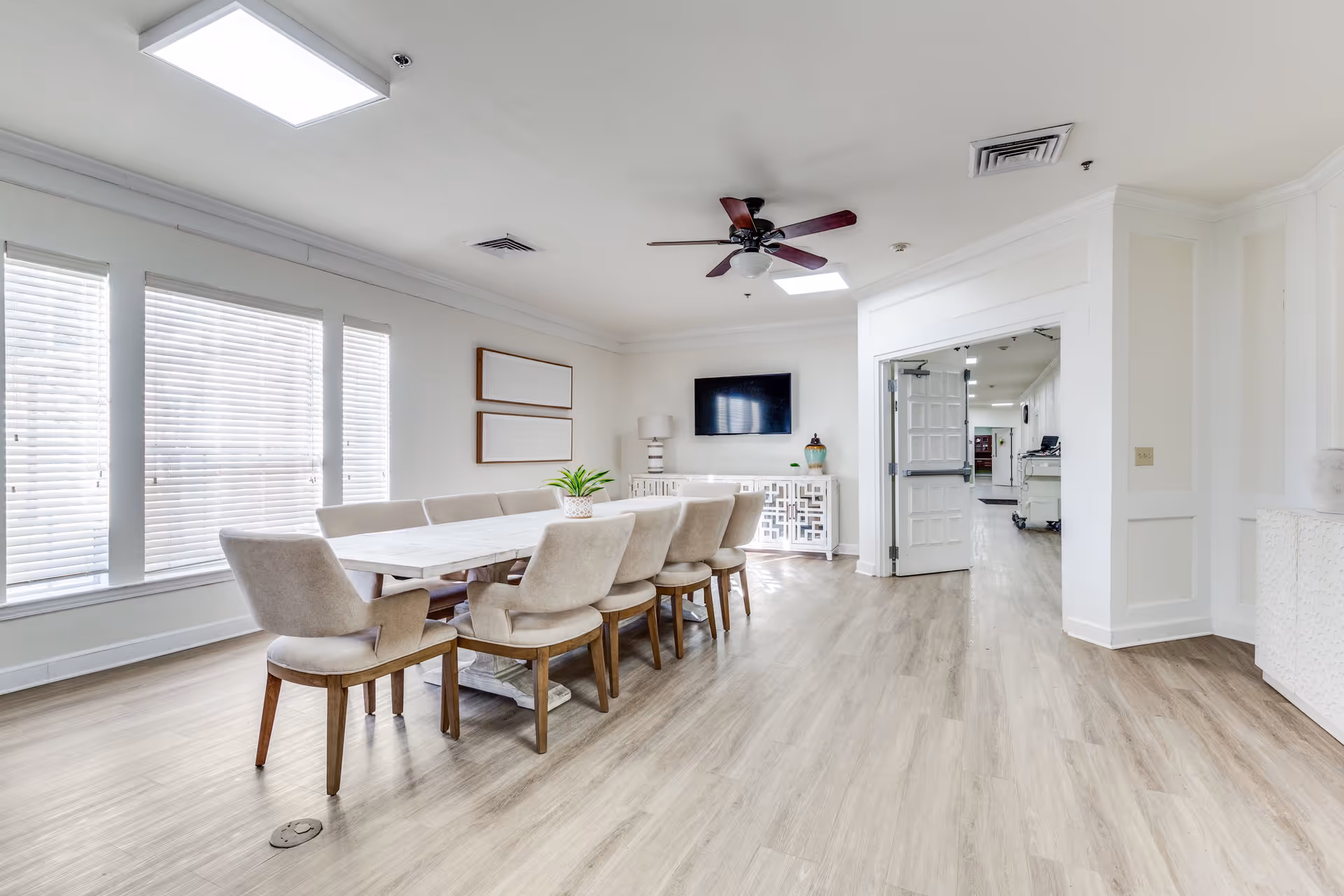 A bright and spacious dining area with a long white table surrounded by eight beige upholstered chairs. The room has large windows with white blinds, light wood flooring, white walls, a ceiling fan with dark wooden blades, and a flat-screen TV mounted on the far wall above a decorative white cabinet. There are two empty picture frames on the wall and a small green plant on the table.